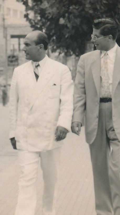 Two men walking together on a street, dressed in vintage suits, one in a white suit and the other in a light gray suit.
